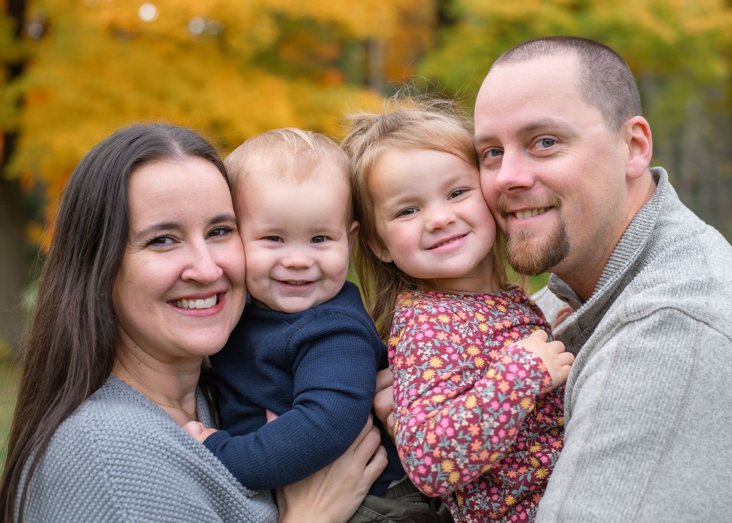 Family of four share a close embrace while smiling at camera with autumn colors behind them.