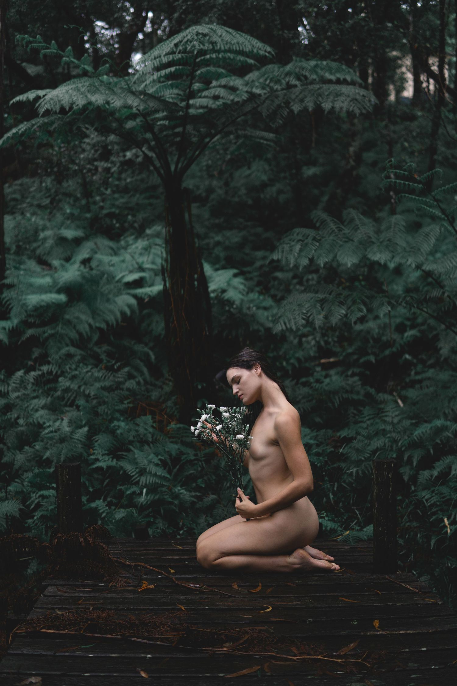 Artistic nude figure holding flowers amid lush green tree ferns in a moody forest setting.