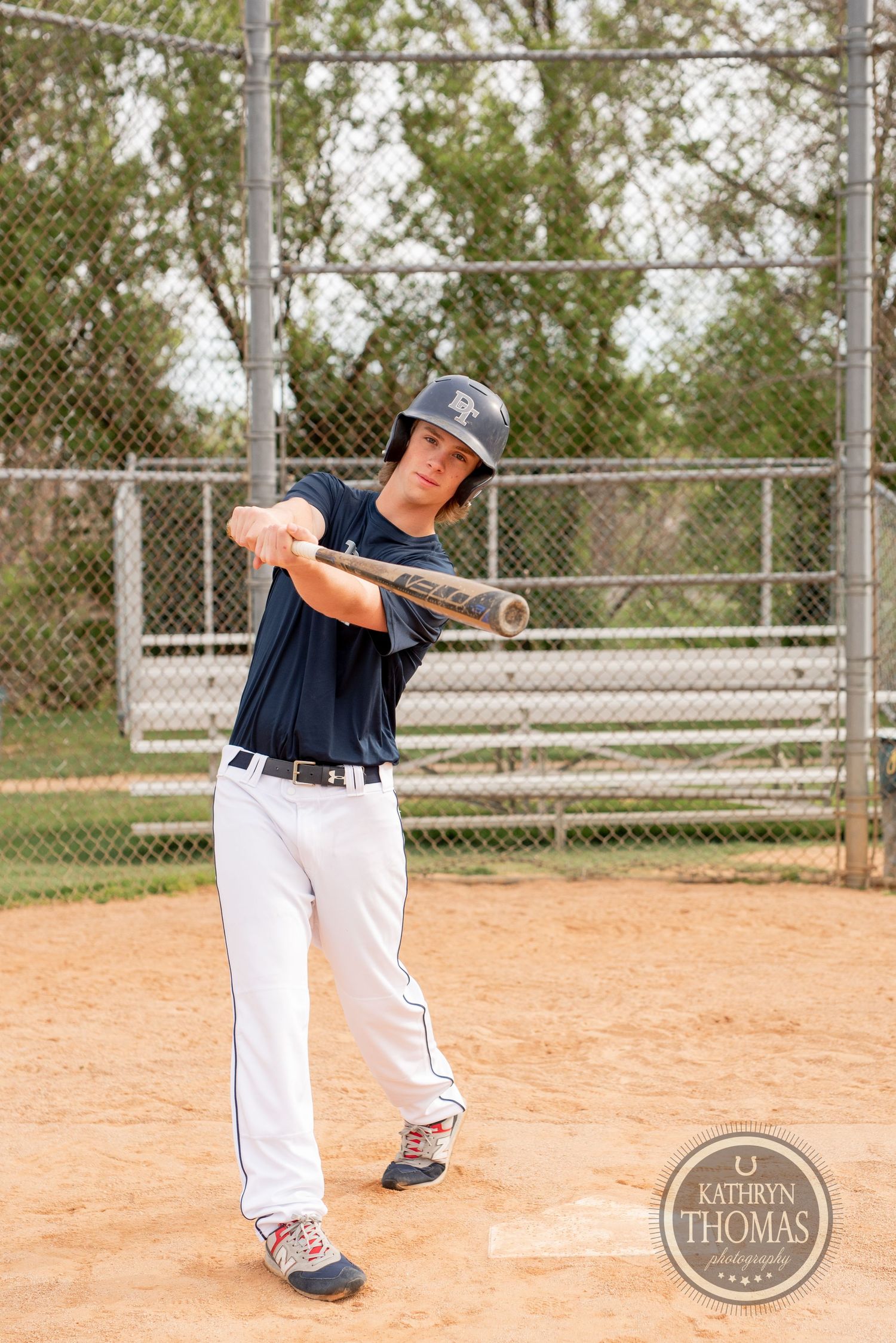 Baseball player in navy uniform demonstrates batting stance on dirt field with chain-link fence in background.