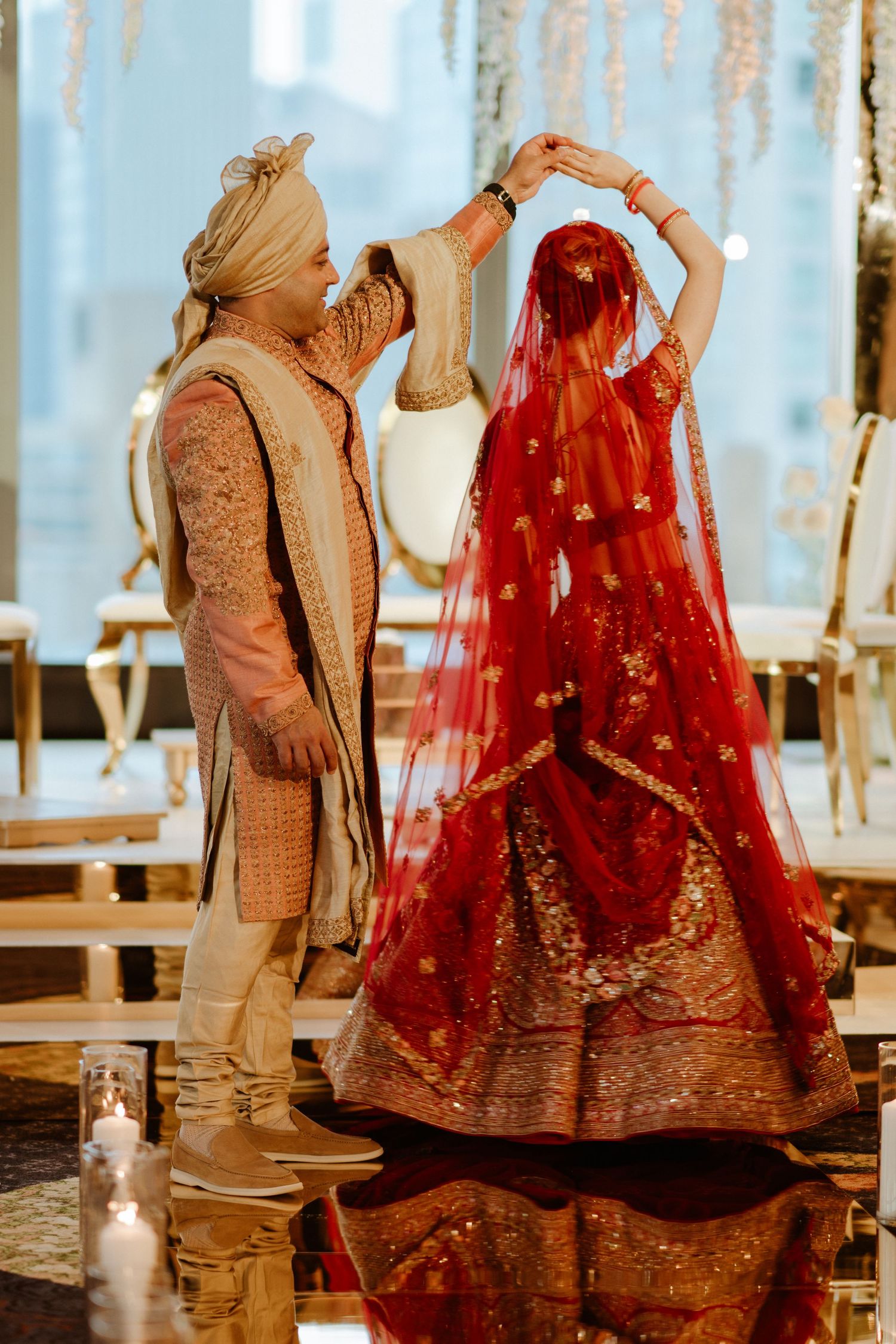 Traditional Indian wedding couple in beige and red outfits photographed against reflective floor.