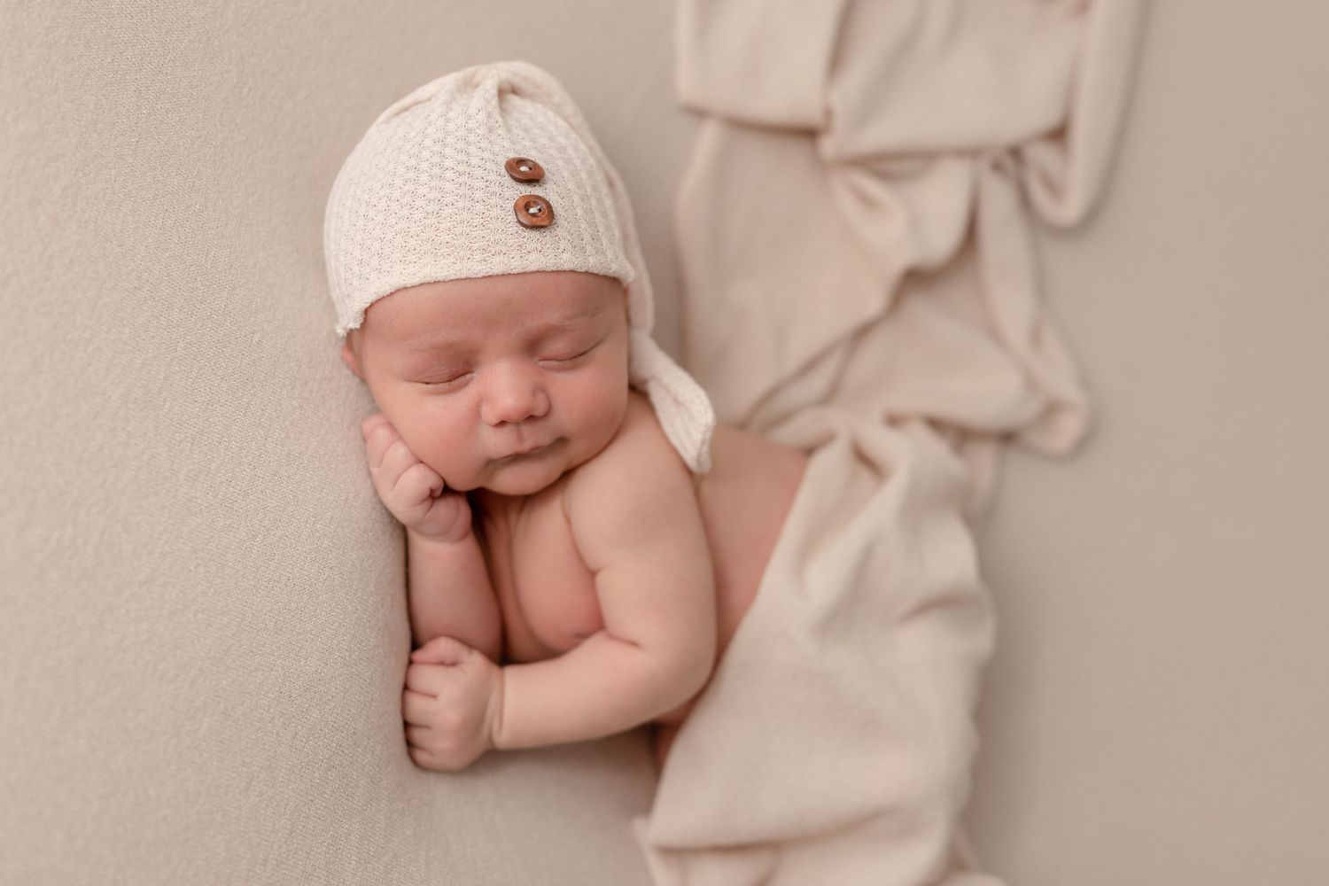 Sleeping newborn wearing white knit hat resting on soft beige fabric.