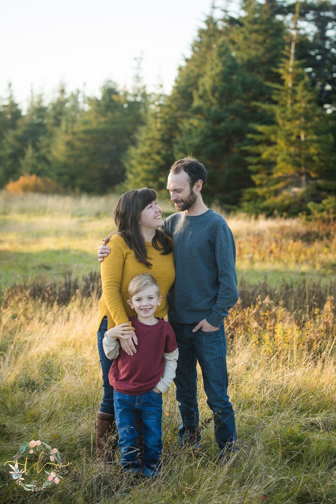 Family shares tender moments together in a golden field during sunset with evergreen trees in the background.