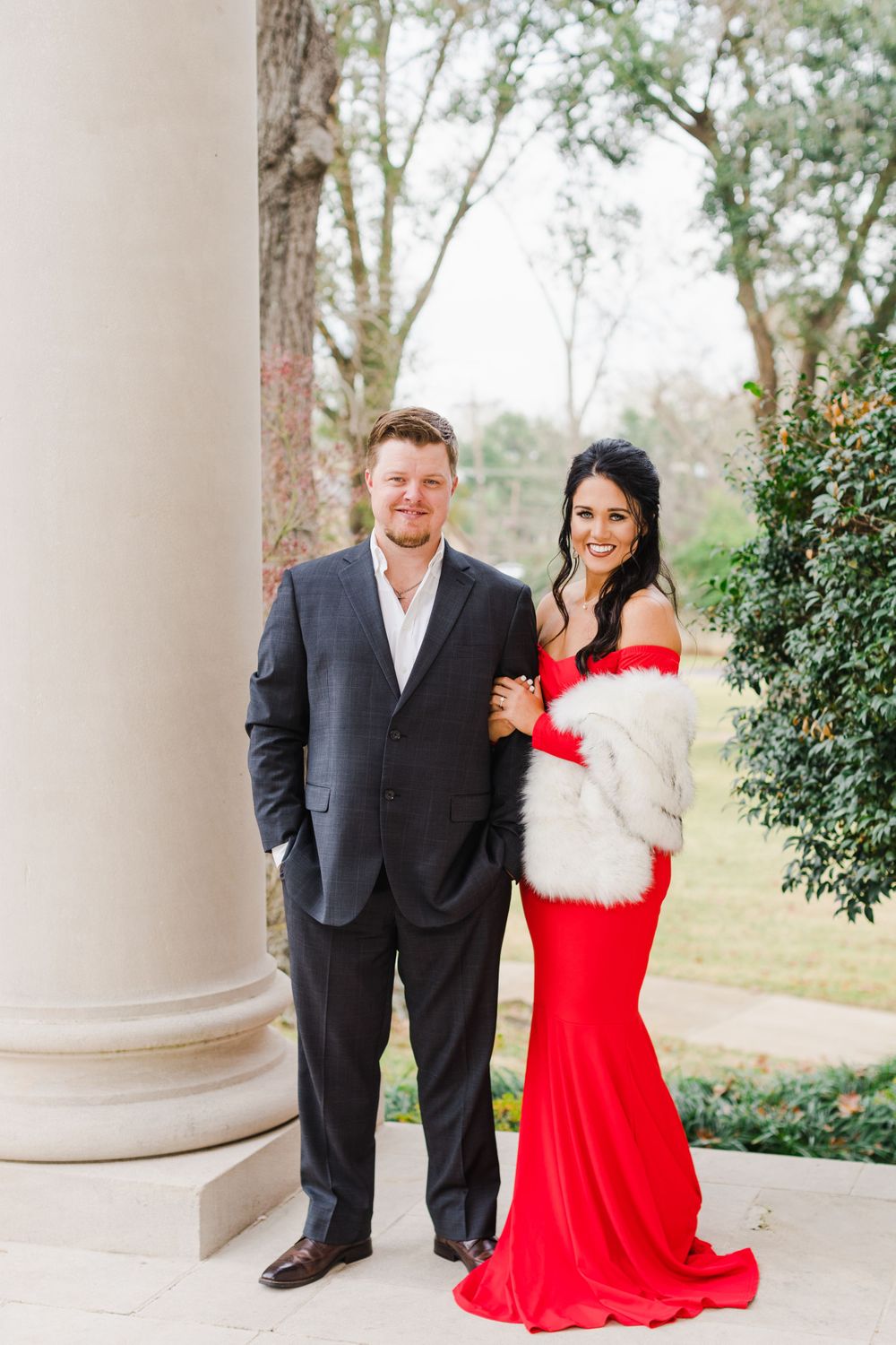 Couple poses elegantly in formal attire with red dress and dark suit near white columns.