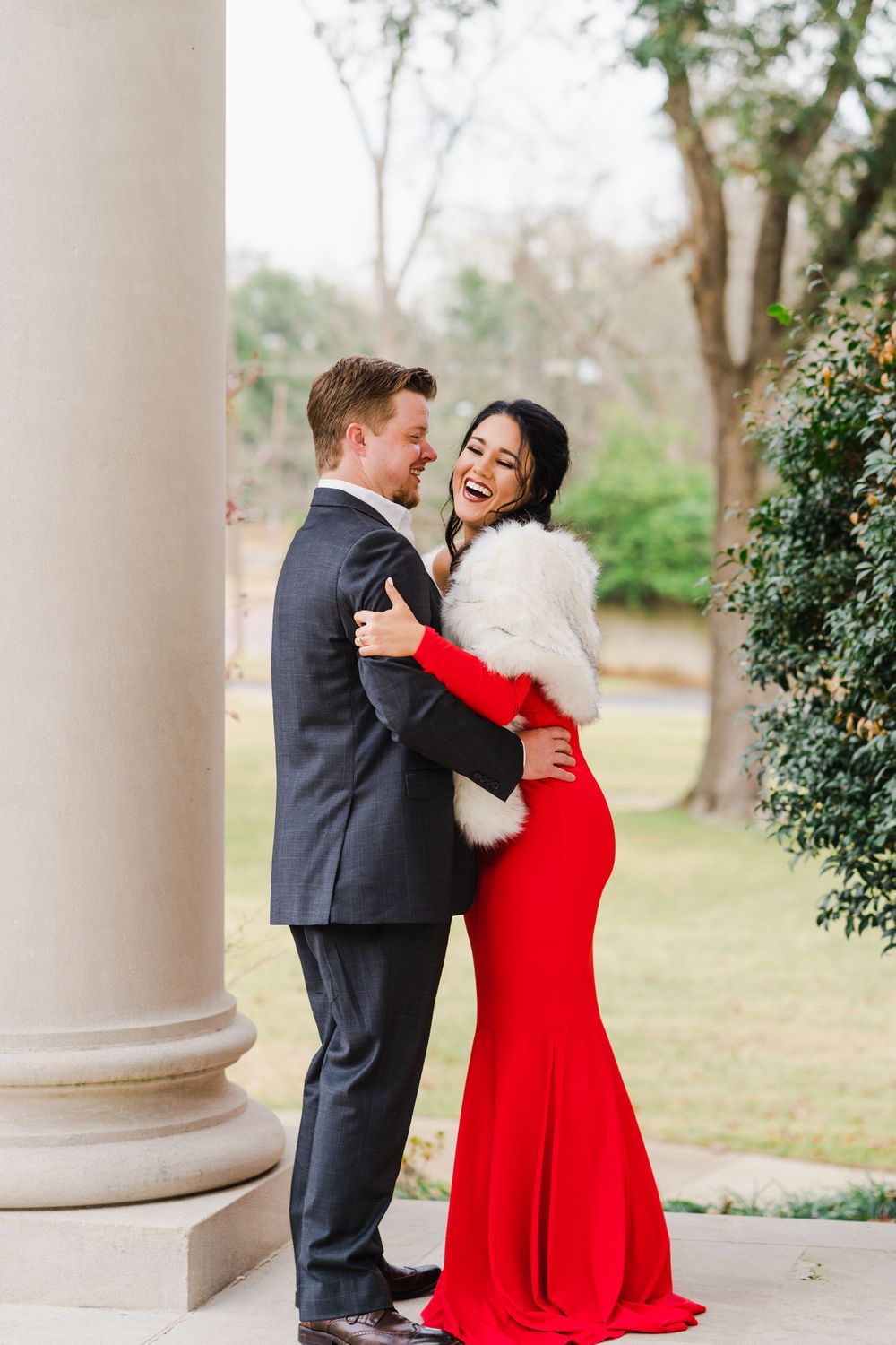 Couple shares intimate moment wearing formal attire with red gown and dark suit by classic columns.