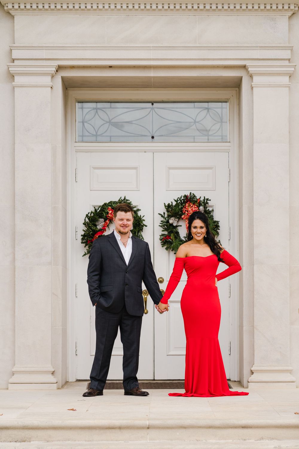 Couple in formal wear poses in front of white door with holiday wreaths.