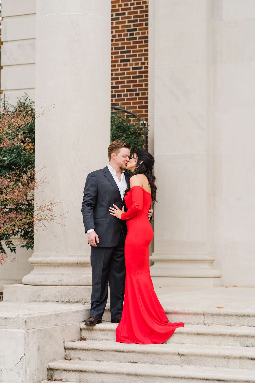 Elegant couple in red dress and dark suit pose on steps by white columns.