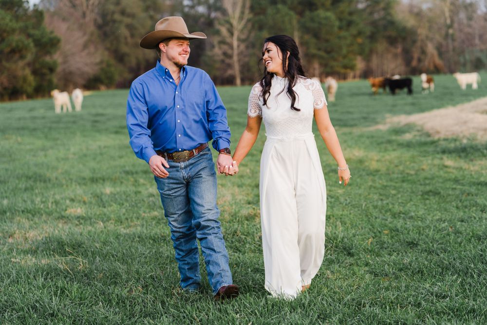Couple walking hand in hand through green pasture with cattle in distance.