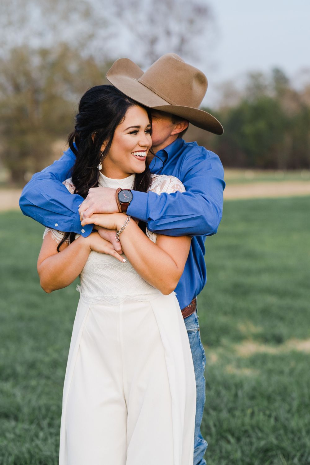 Joyful embrace in countryside setting with bright blue shirt and white jumpsuit.