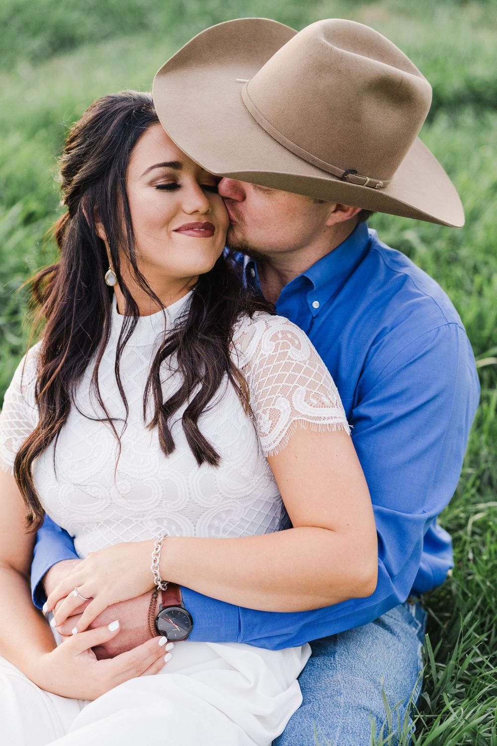 A romantic couple embraces in a grassy field with the person wearing a cowboy hat and blue shirt.