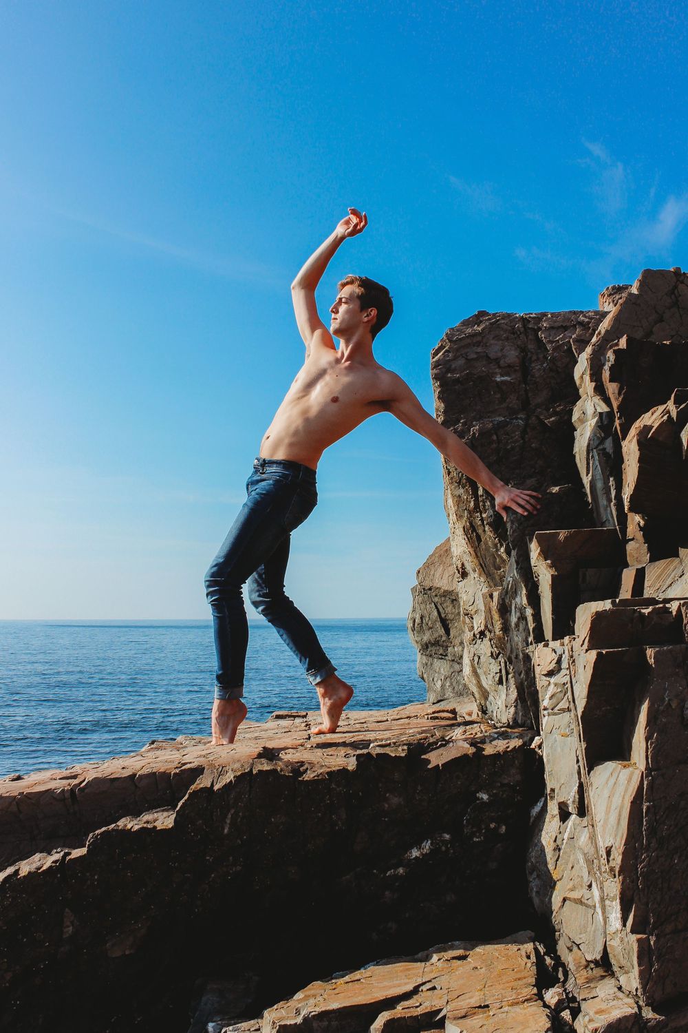 Person striking dramatic pose on rocky ocean cliff edge at sunset.