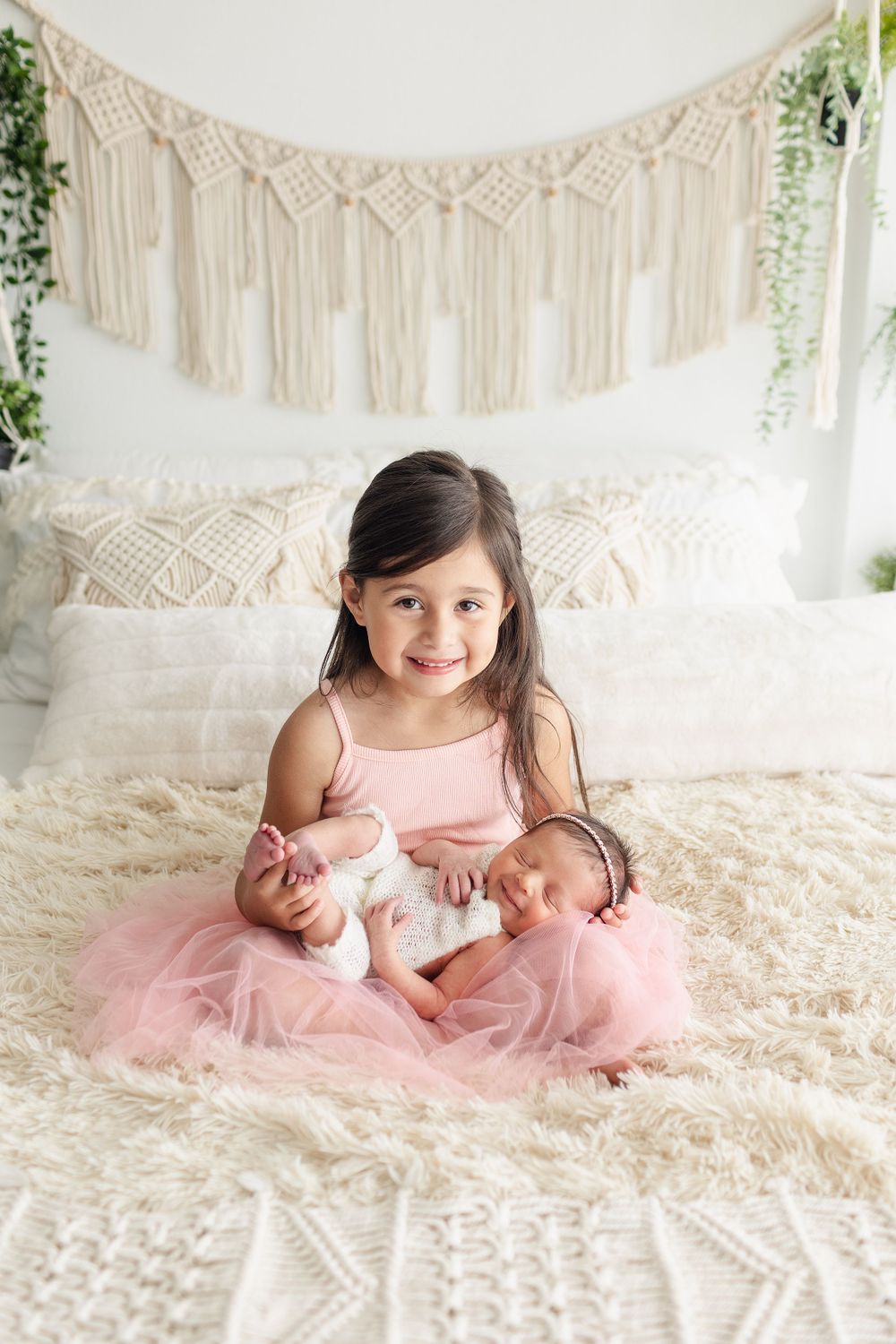 Joyful big sister in pink tulle dress with sleeping newborn on cream bedding.
