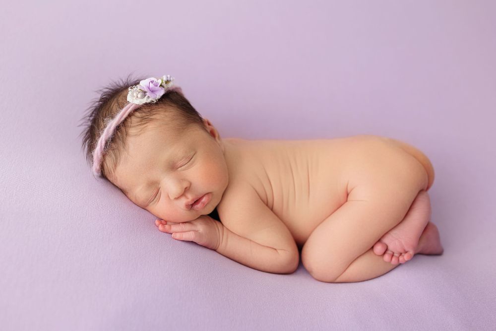 Sleeping newborn baby wearing floral headband posed on purple background.