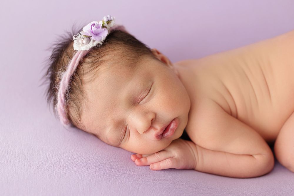 Sleeping newborn baby wearing pink floral headband resting on purple backdrop.