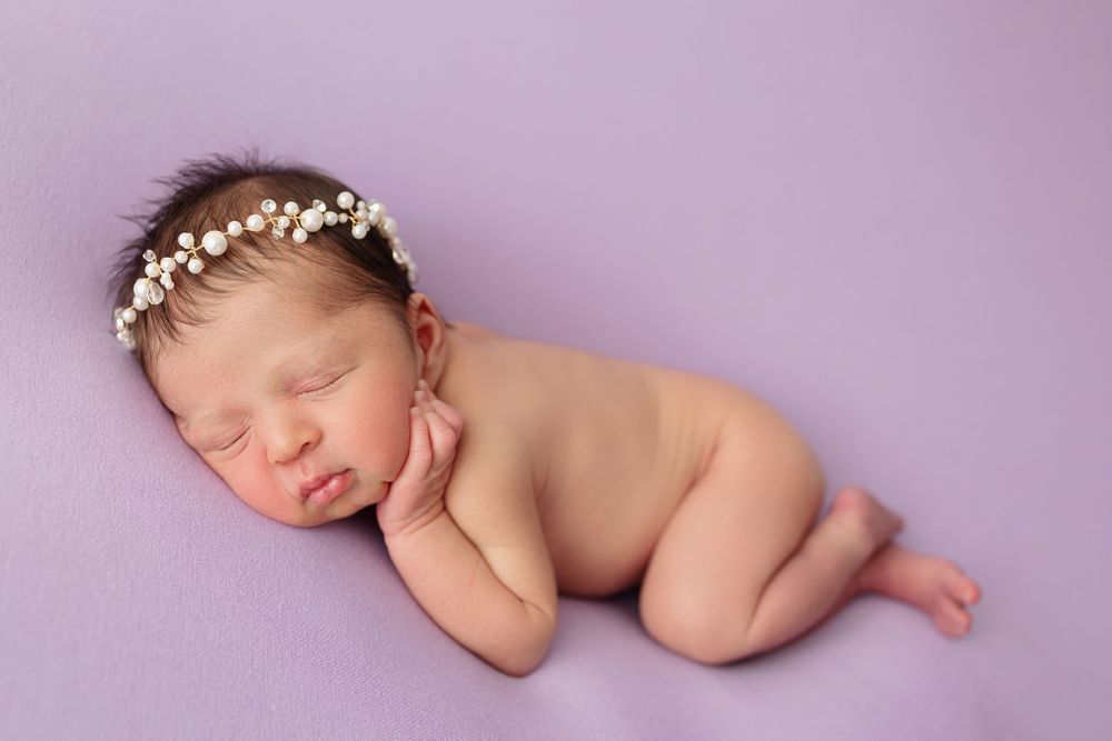 Newborn baby wearing delicate flower crown sleeping peacefully on purple backdrop.