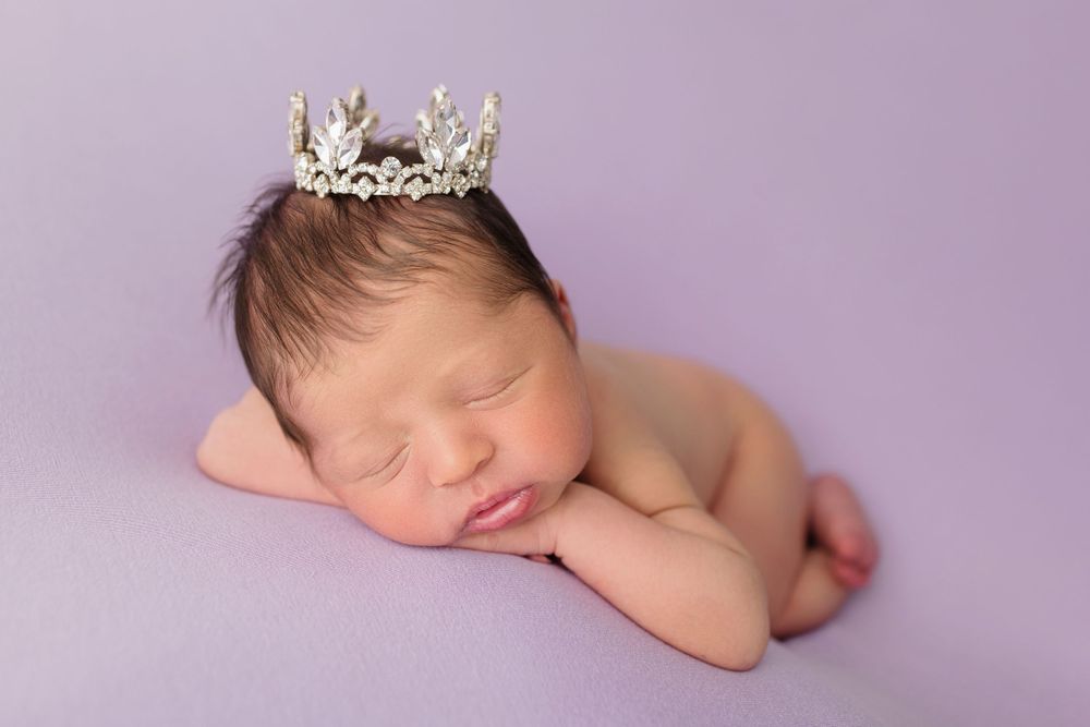 Sleeping newborn wearing silver crown posed on purple background.