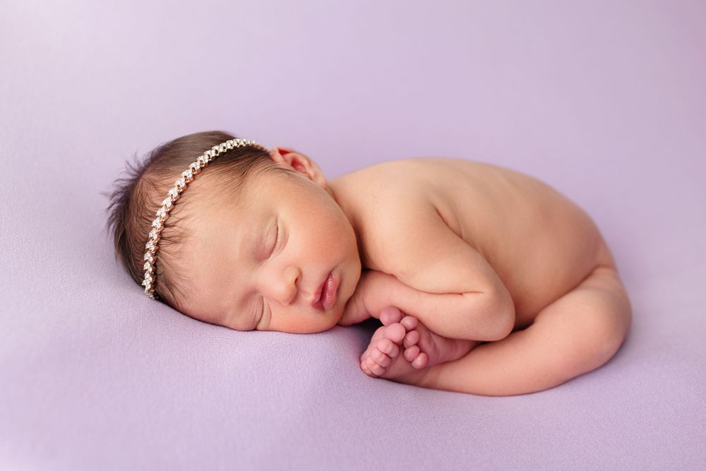 Peaceful newborn with pearl headband sleeping on purple backdrop.