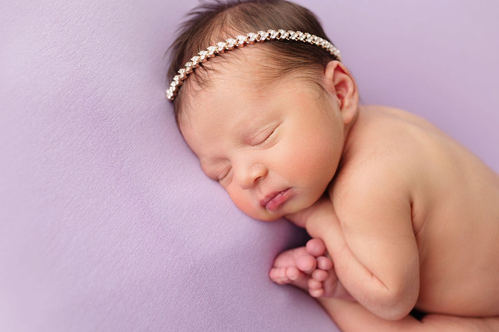 Sleeping newborn wearing delicate pearl headband on purple background.