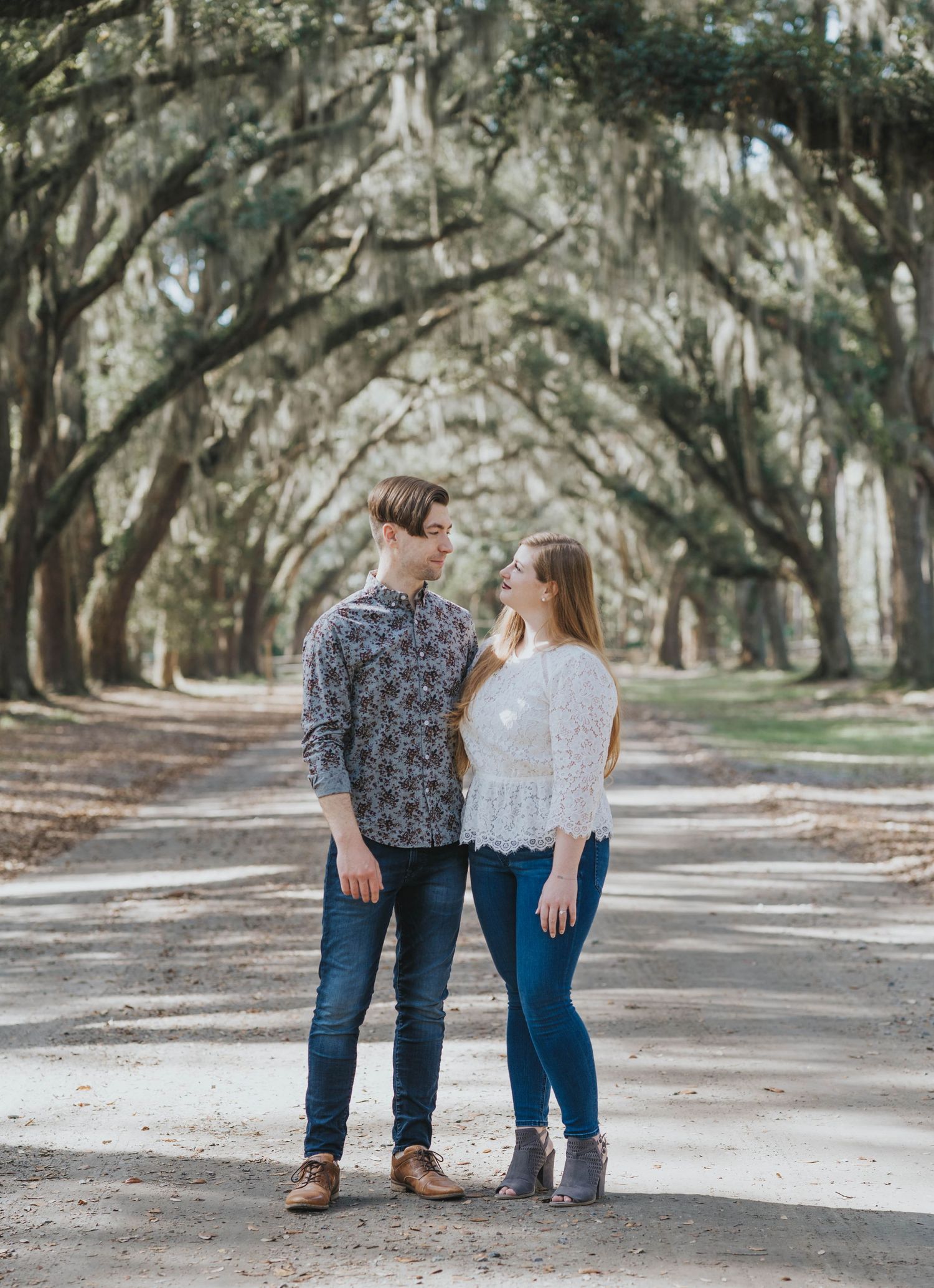 A couple stands together on a tree-lined path with Spanish moss hanging from oak branches overhead.
