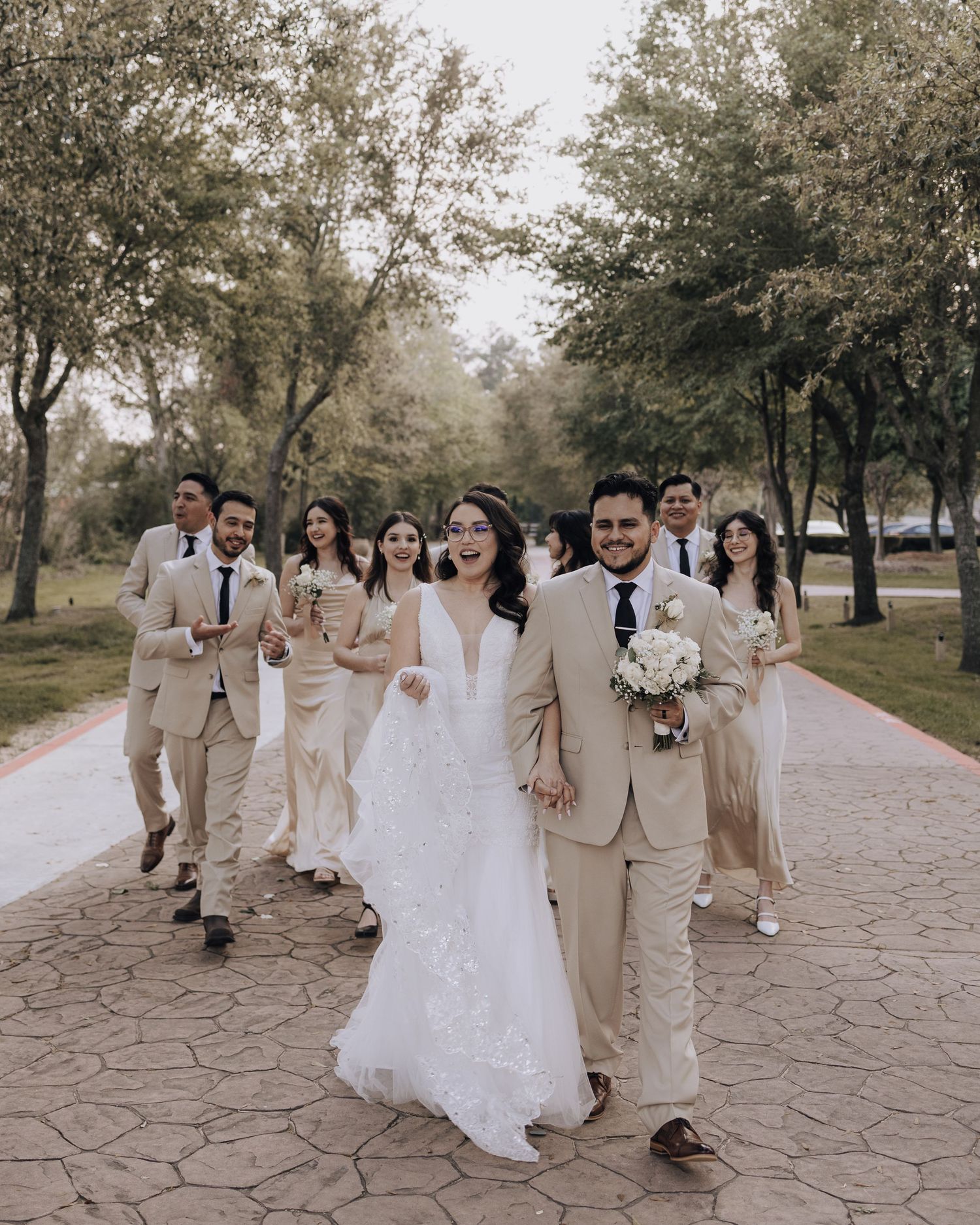 Wedding party walks down stone path amid trees wearing coordinated beige and white attire.