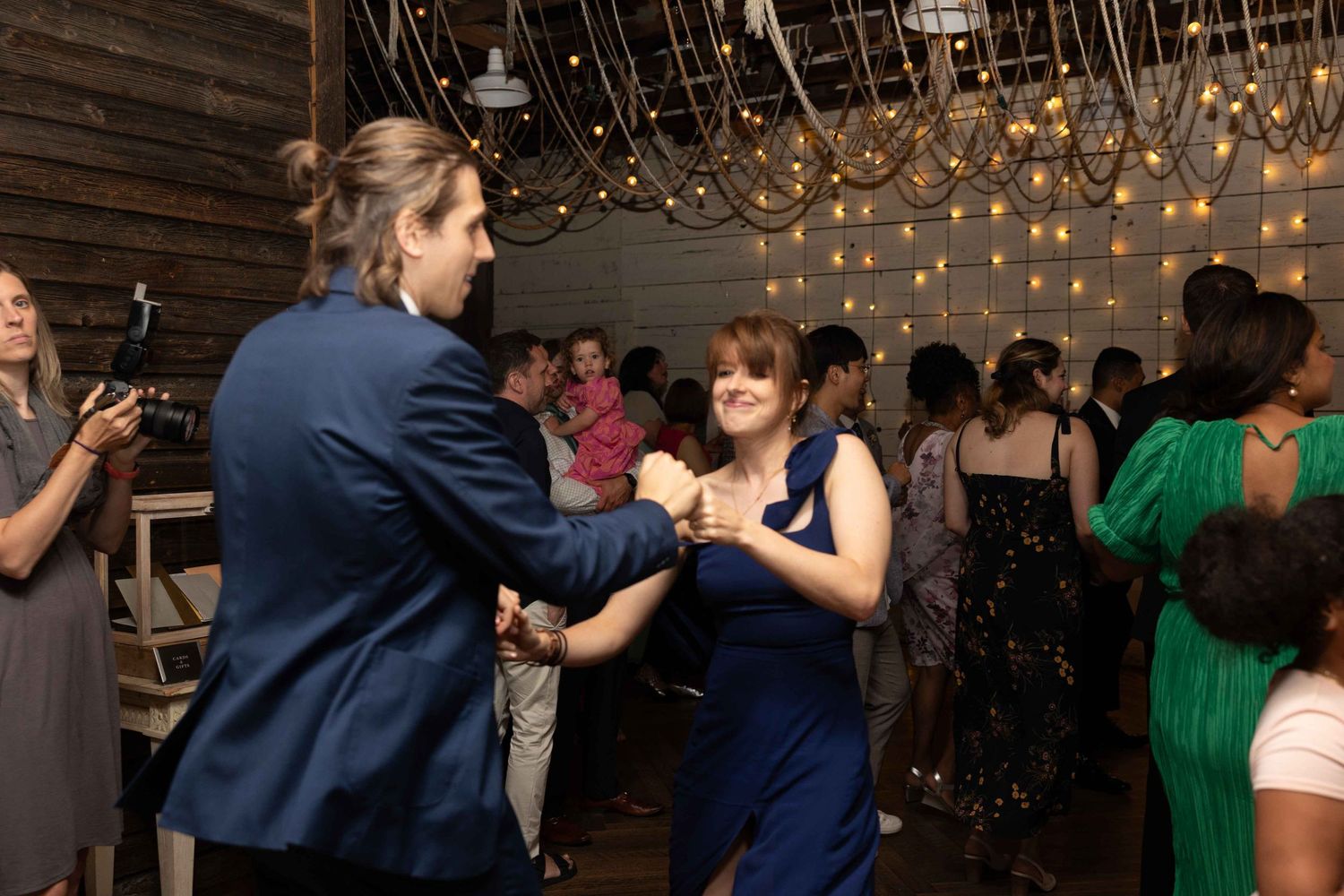 Couples dancing in a rustic venue decorated with string lights hanging from wooden beams.