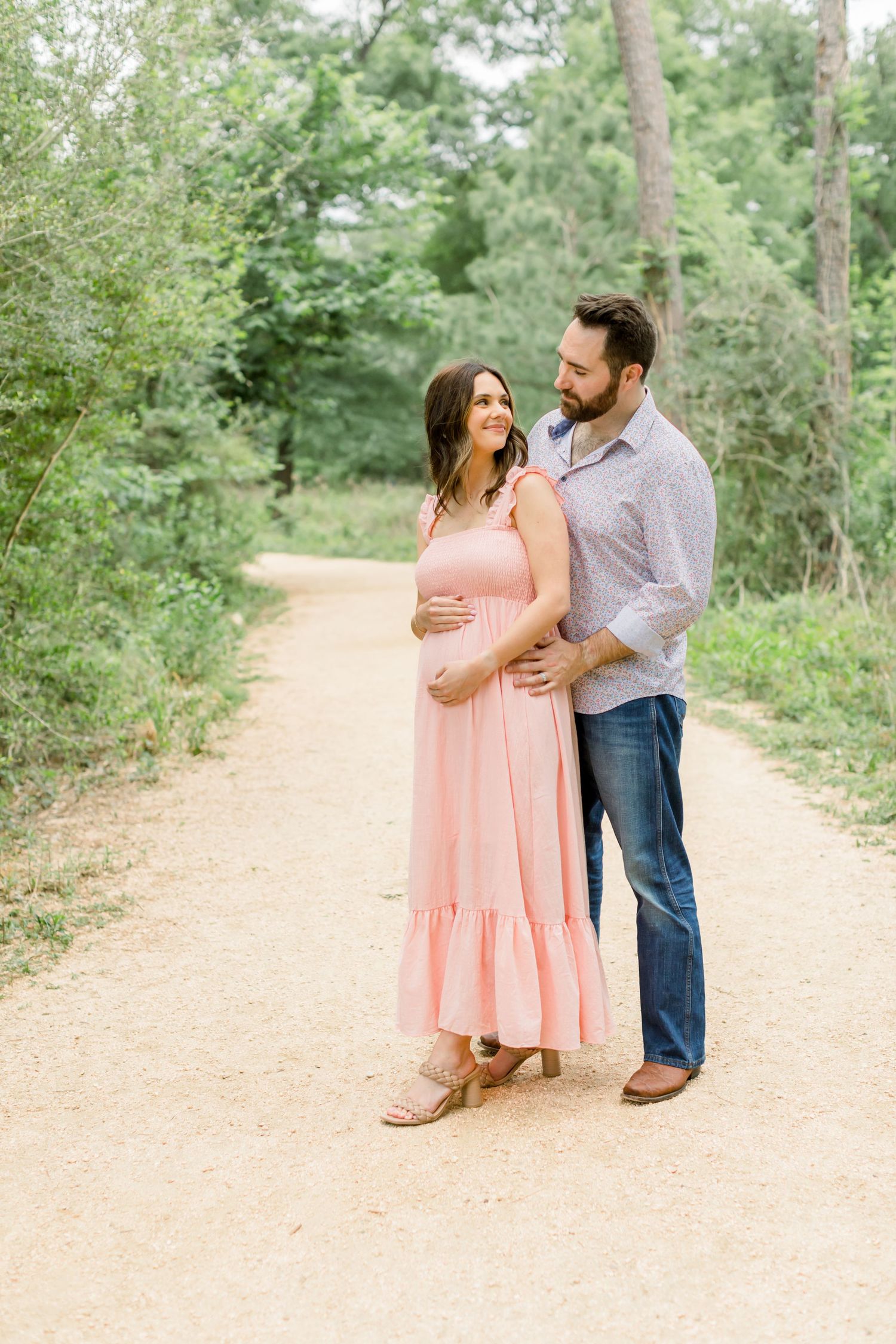 A couple stands together on a nature trail surrounded by lush greenery, with the woman wearing a flowing pink dress.