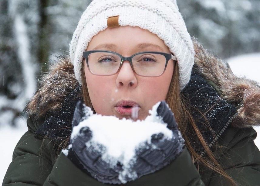 Person in winter hat and glasses blowing snow from gloved hands.
