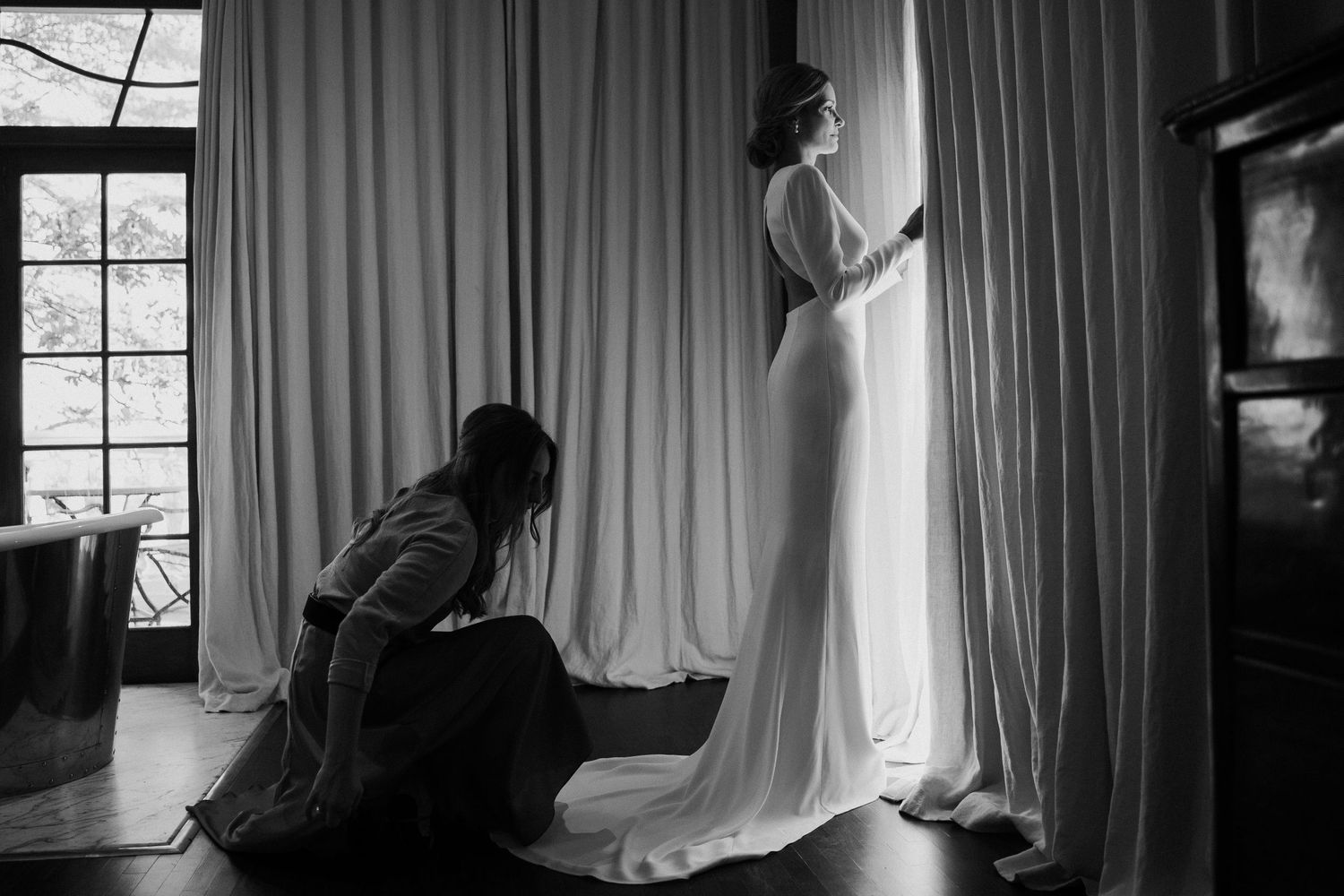 Wedding dress preparation scene with flowing white gown against window light and curtains.