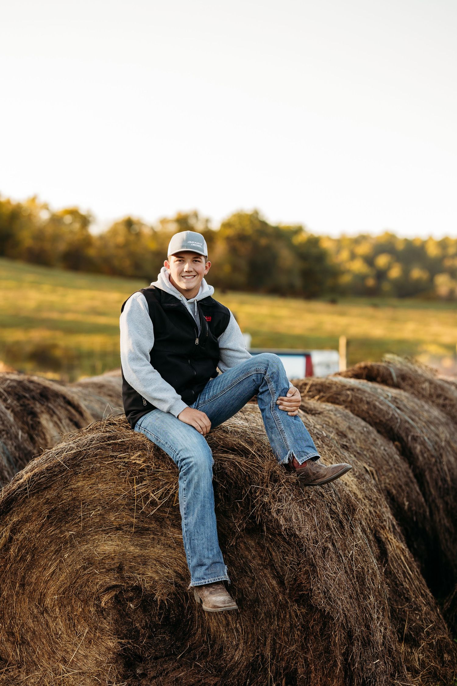 A casual portrait of someone sitting on hay bales at sunset wearing jeans and a baseball cap.
