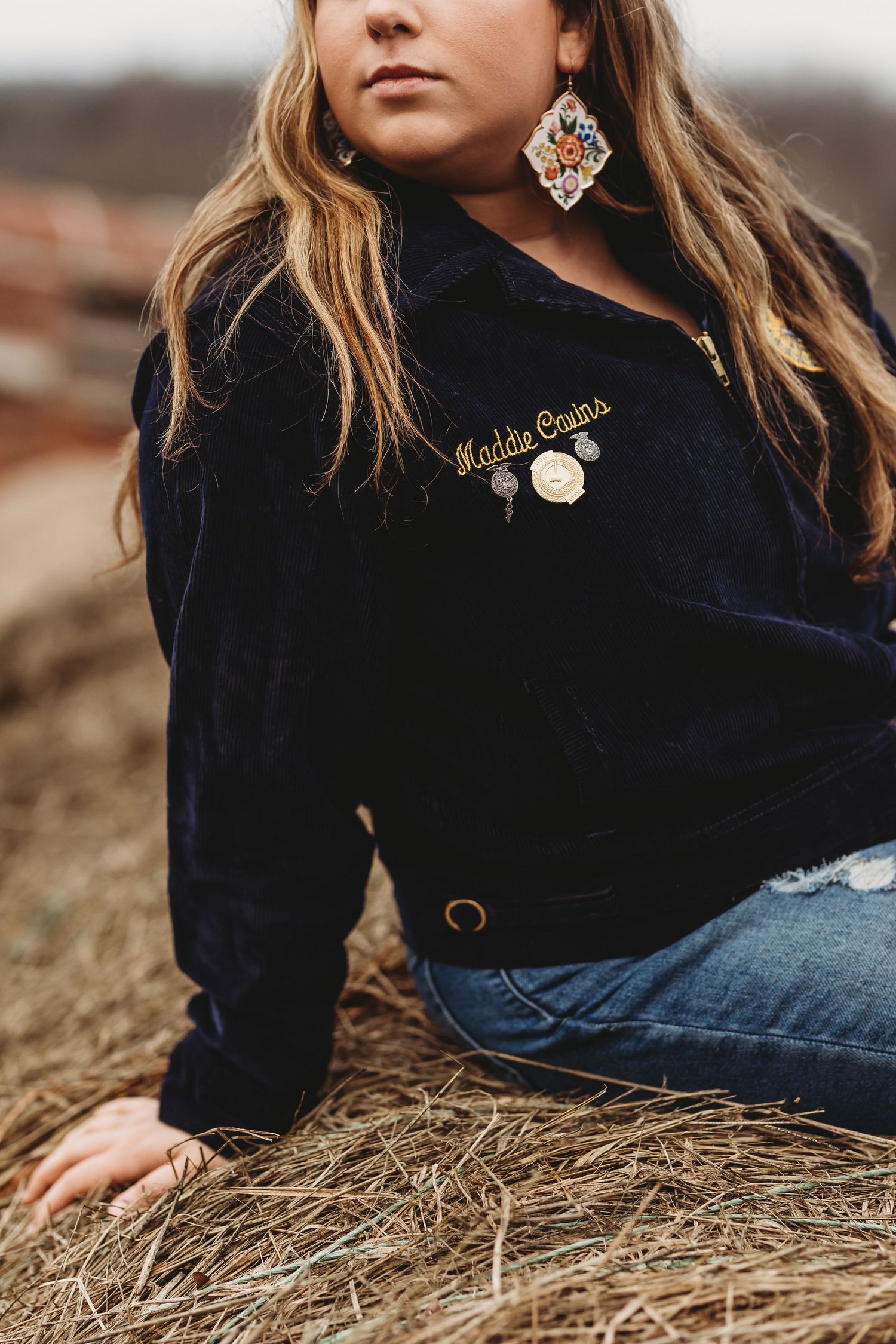 Close-up detail of FFA jacket with pins and decorative earring against rural background.