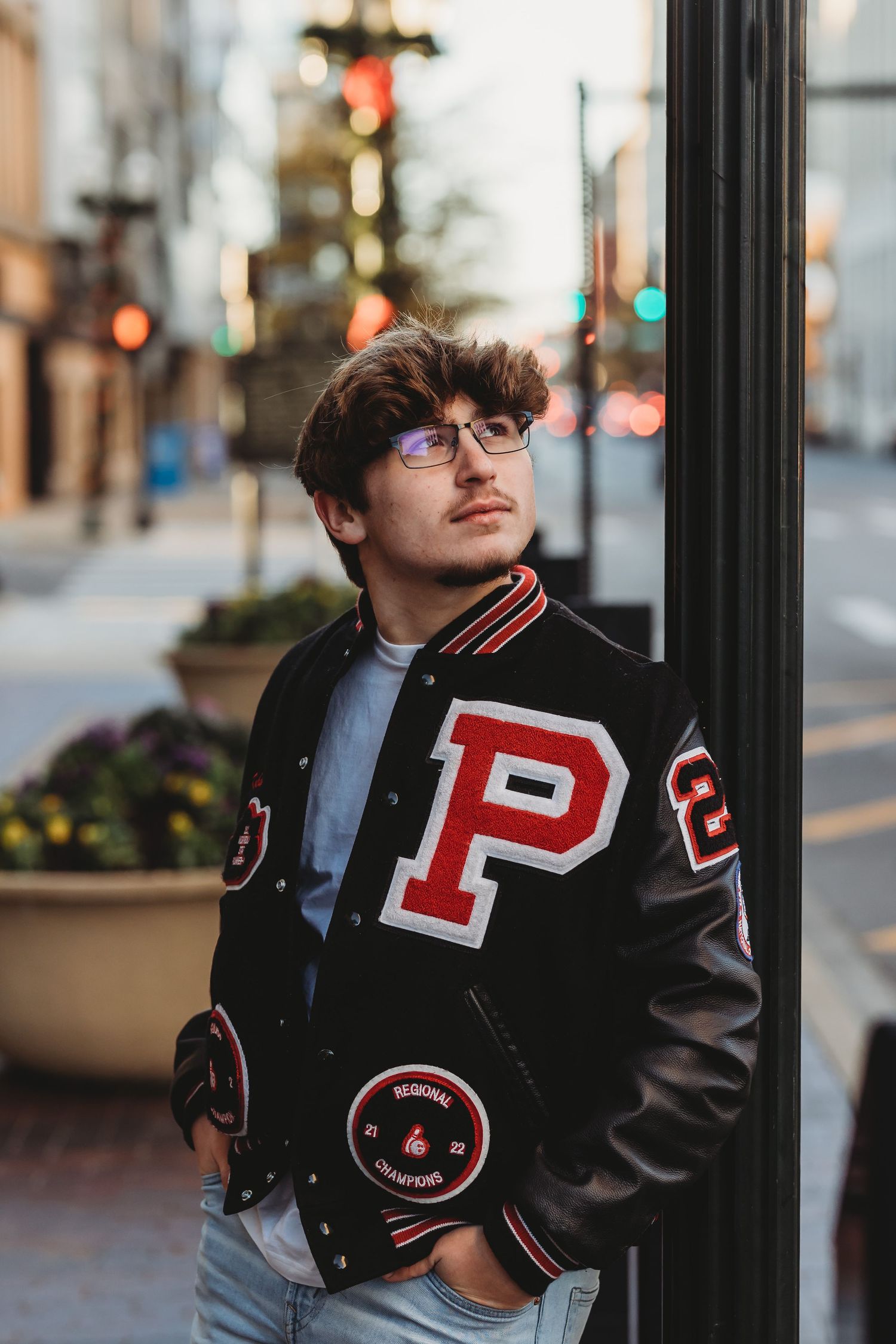 Student in varsity letterman jacket poses against city street pole at dusk.