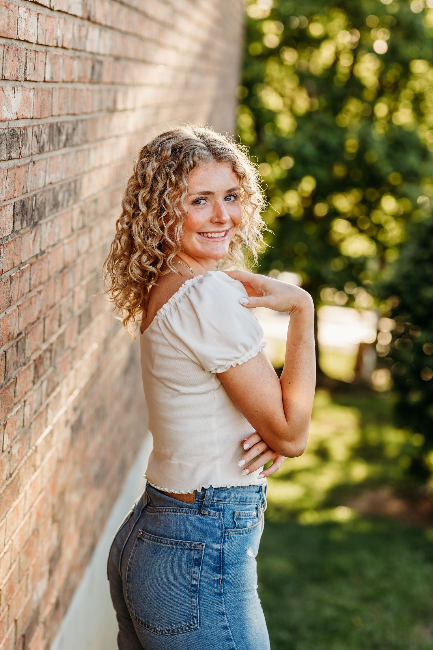 A casual portrait against a brick wall featuring curly blonde hair, white top and jeans in natural lighting.