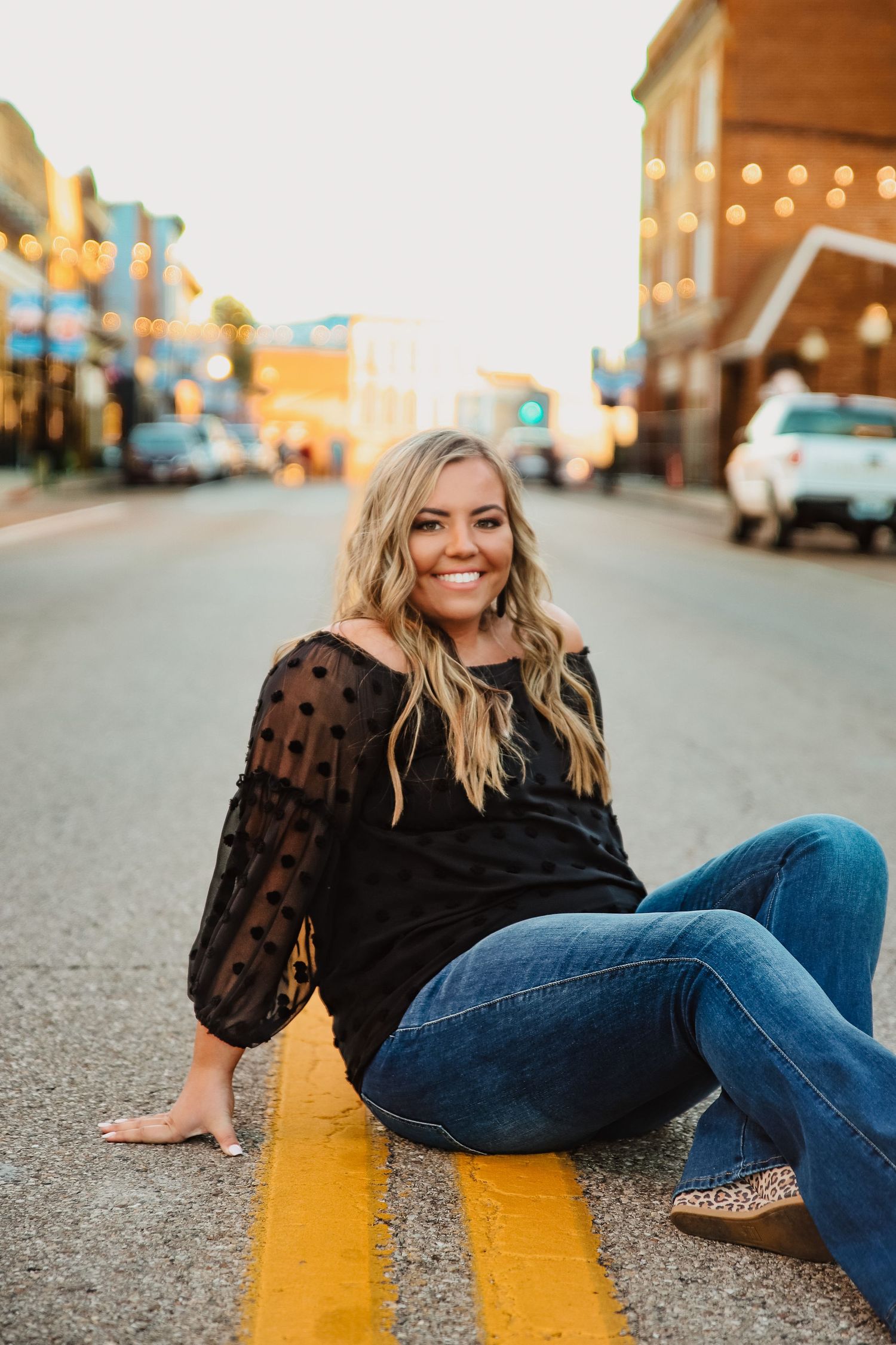 Woman in polka dot blouse sits on yellow street line at dusk with city lights behind her.