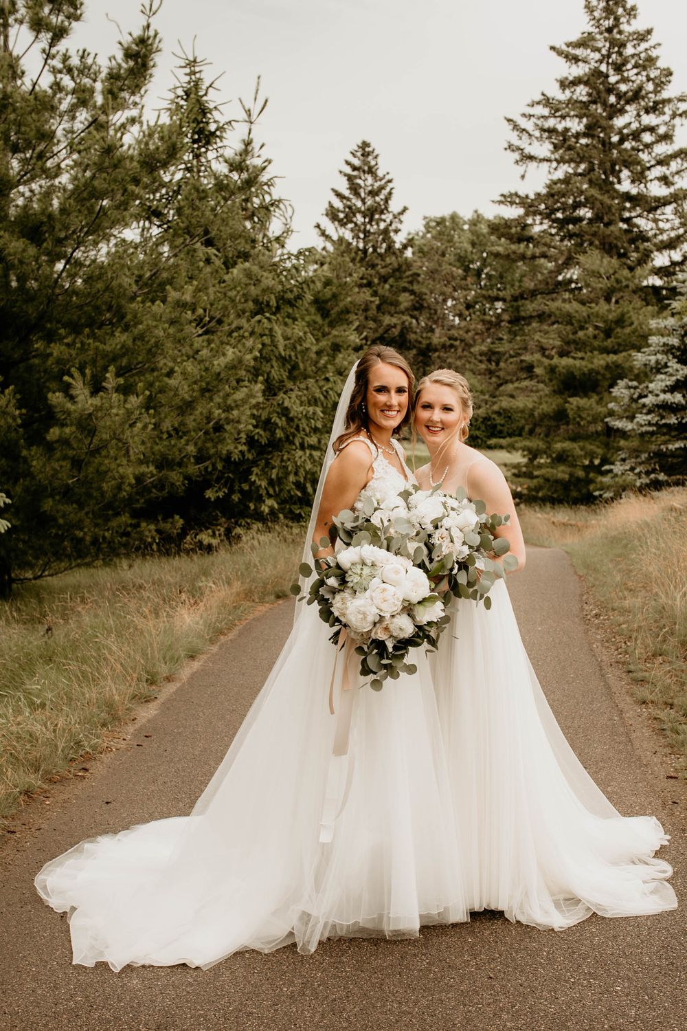 Two brides in elegant white wedding dresses posing together on a scenic outdoor pathway.