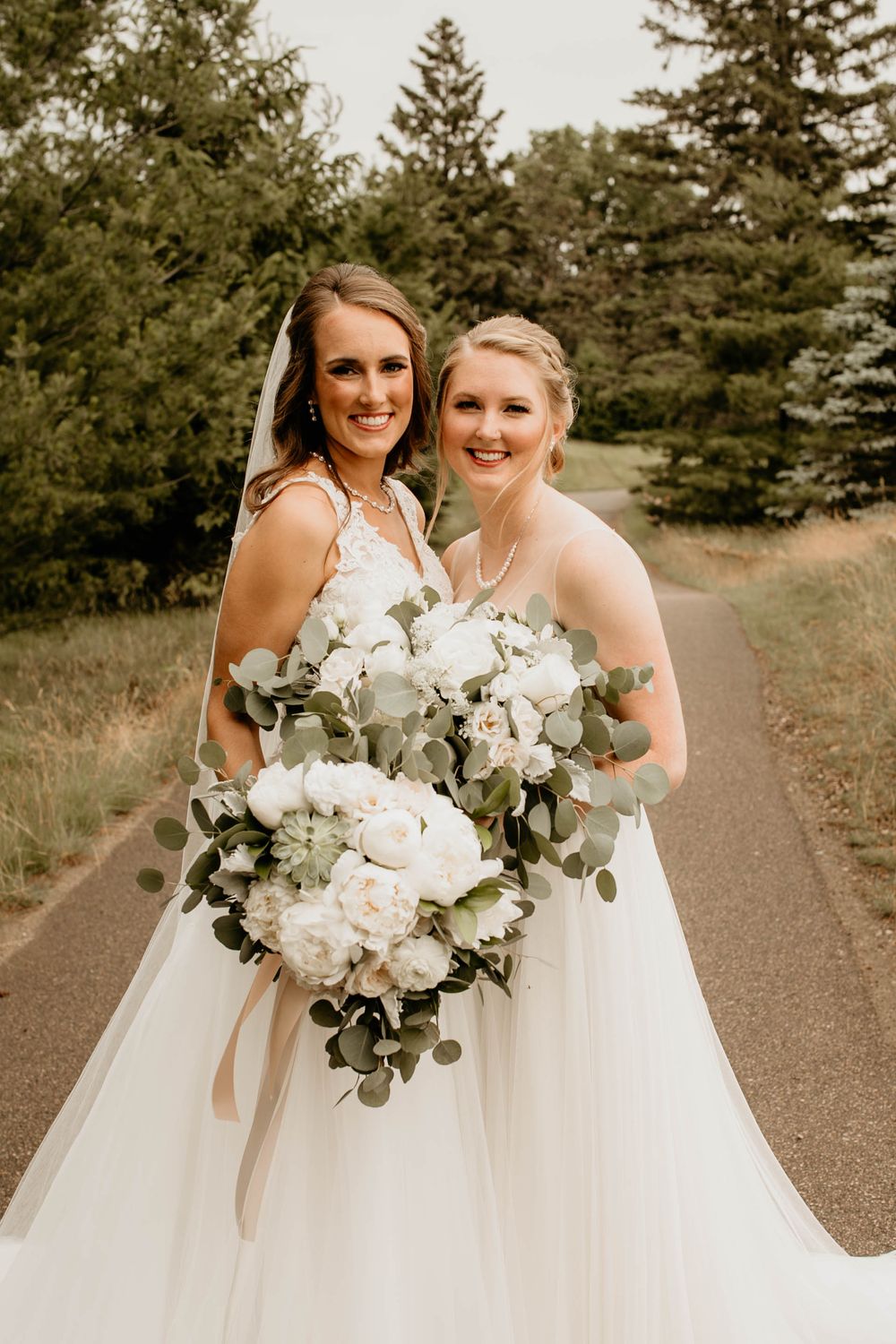 Two brides in white gowns holding white floral bouquets with eucalyptus on a tree-lined path.