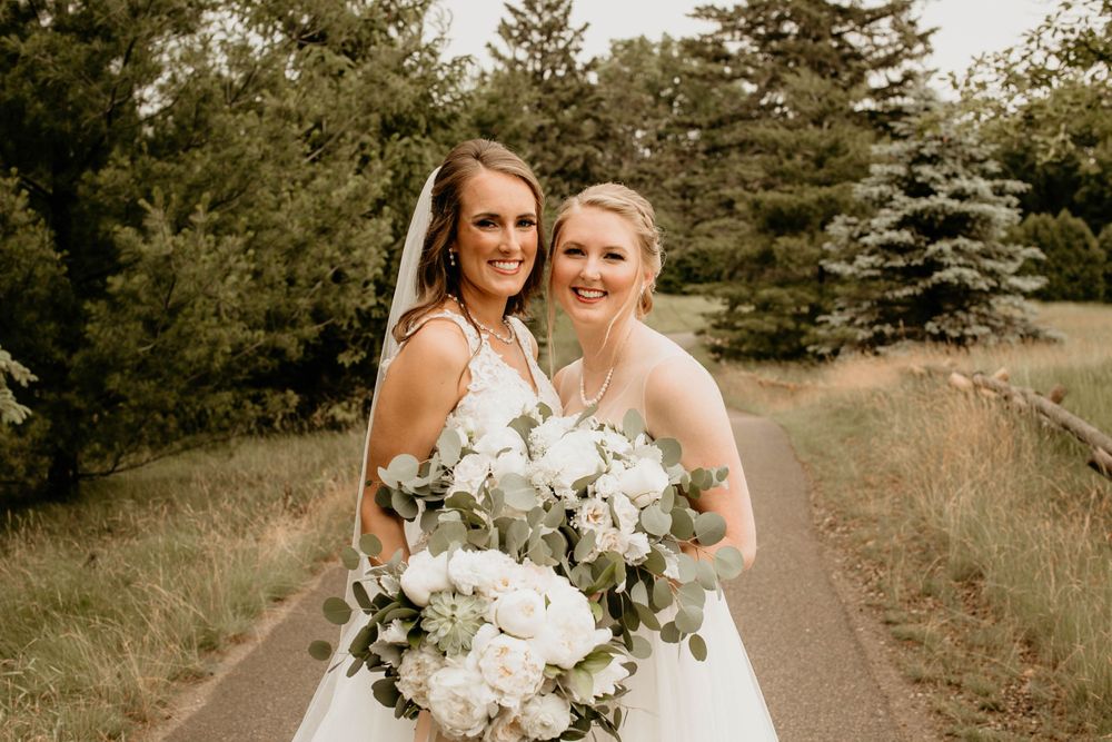 Two smiling brides in white dresses holding white peony and eucalyptus bouquets outdoors.