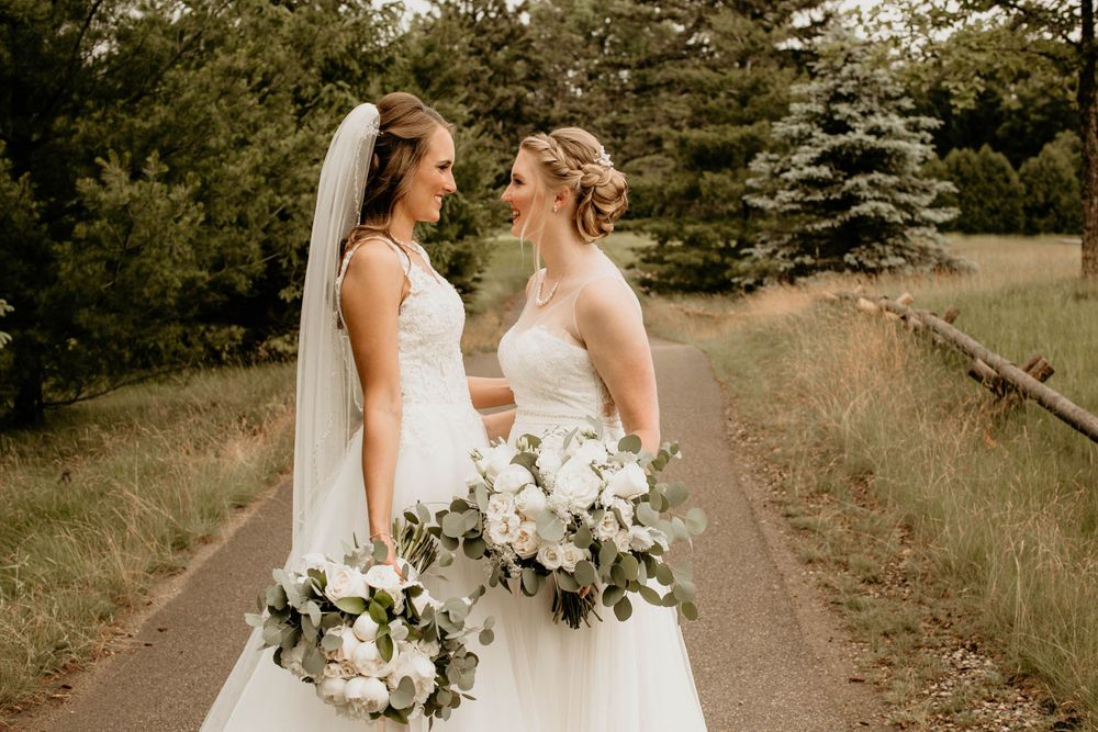 Two brides in white gowns facing each other lovingly on a tree-lined wedding day path.