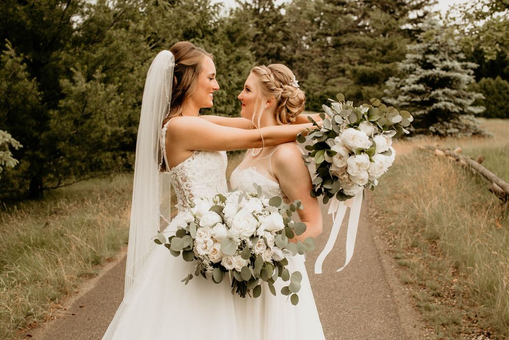 Two brides laughing joyfully together on a path, one lifting her leg in white gowns.