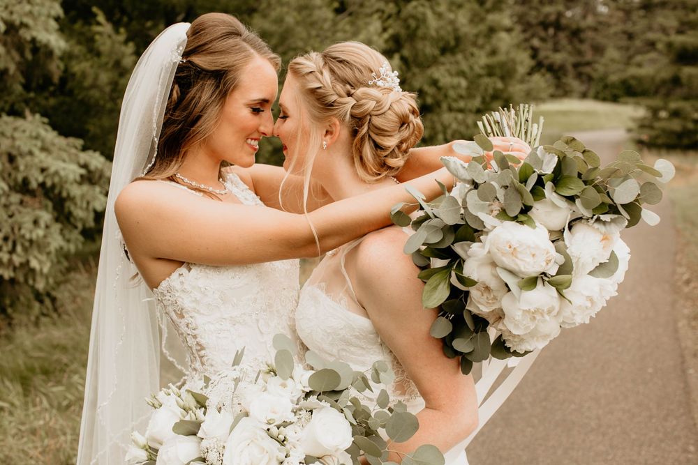 Two brides sharing a tender moment, one wearing a veil, holding white floral bouquets.