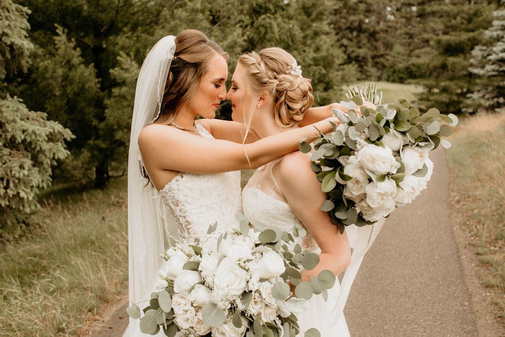 Two brides leaning in close together smiling, holding white and green wedding bouquets.