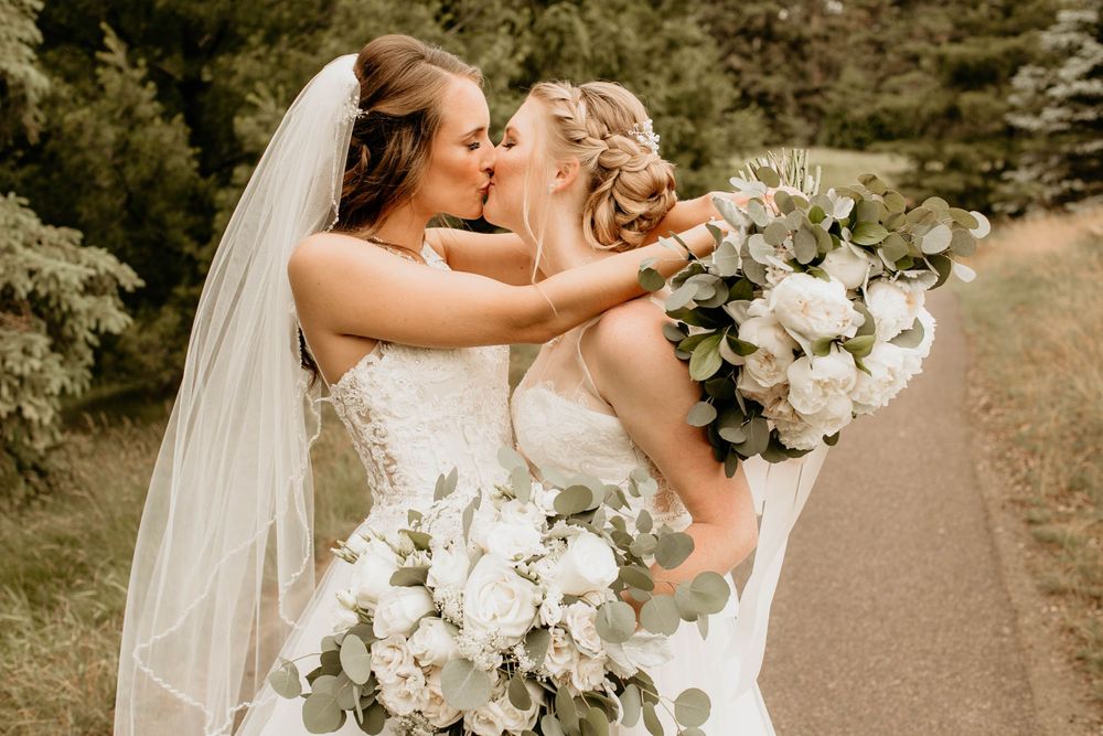 Two brides sharing a kiss on their wedding day while holding white floral bouquets.
