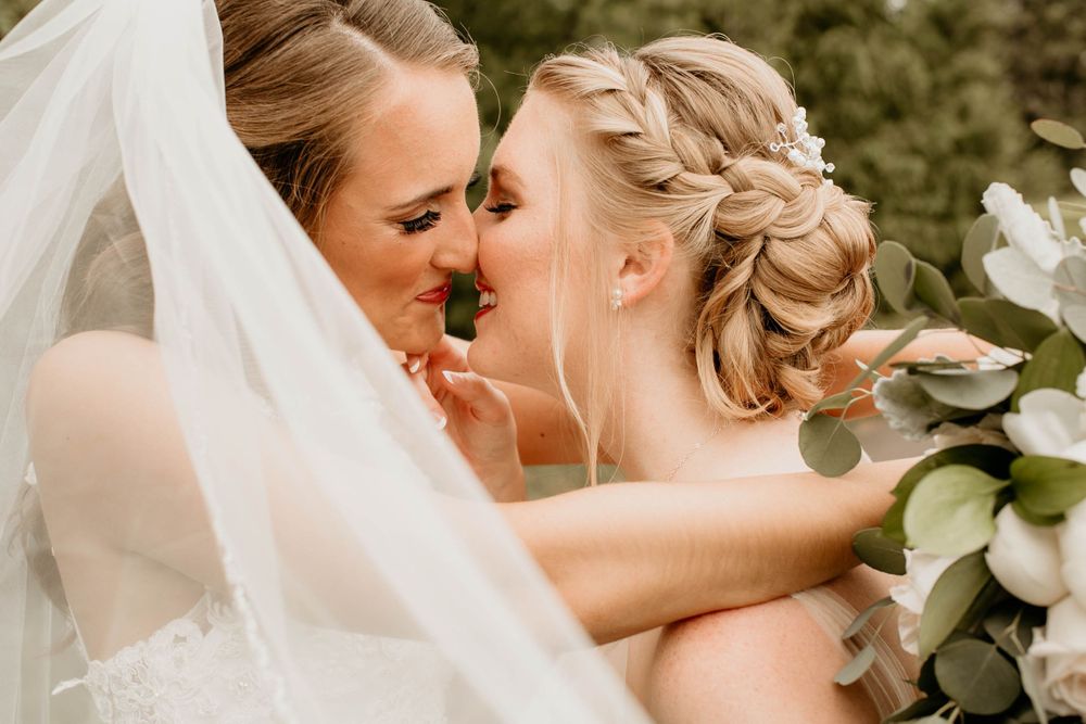 Two brides sharing a tender kiss beneath a white veil with eucalyptus bouquet nearby.