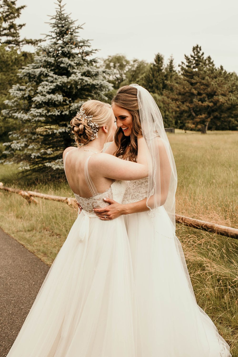 Two brides in white gowns share a tender embrace outdoors, surrounded by lush green trees on their wedding day.