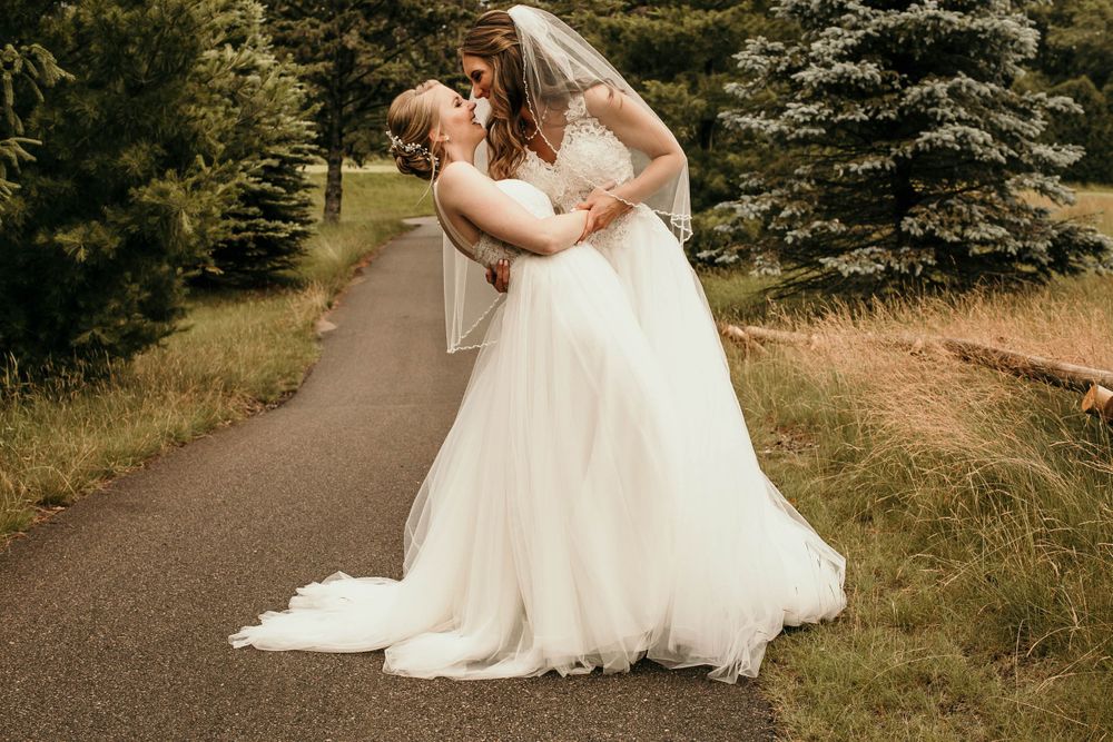 Two brides in elegant white wedding gowns share a romantic kiss on a tree-lined pathway on their wedding day.