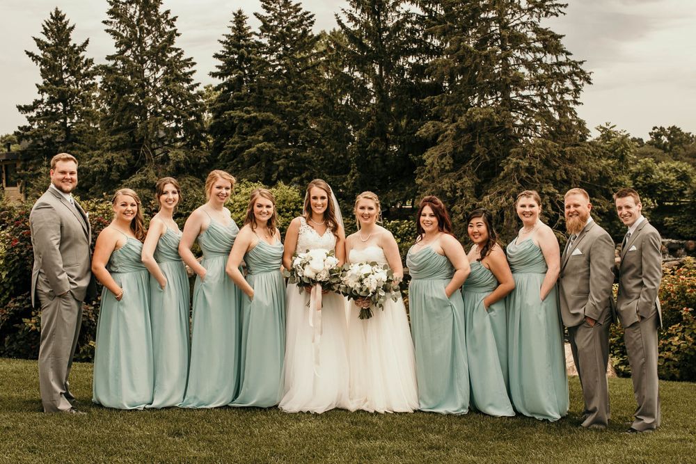 Two brides pose with their wedding party in sage green bridesmaid dresses and grey suits on a lush green lawn.