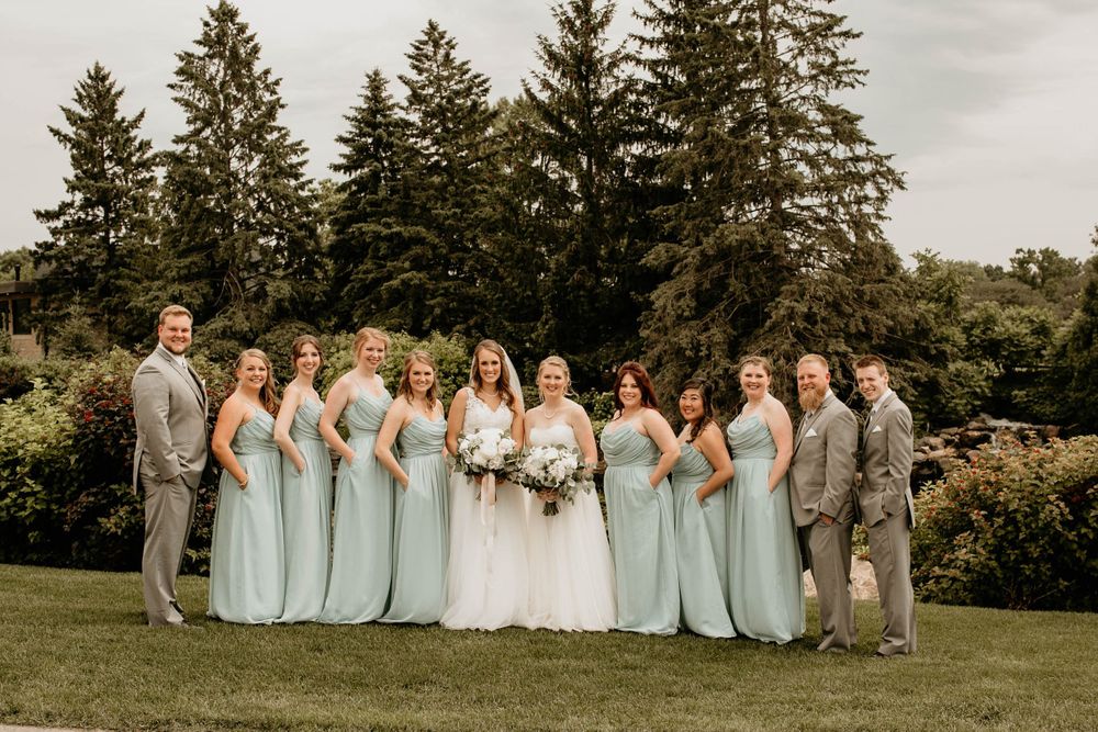 Two brides stand with bridesmaids in mint green gowns and groomsmen in grey suits for a formal wedding party photo.