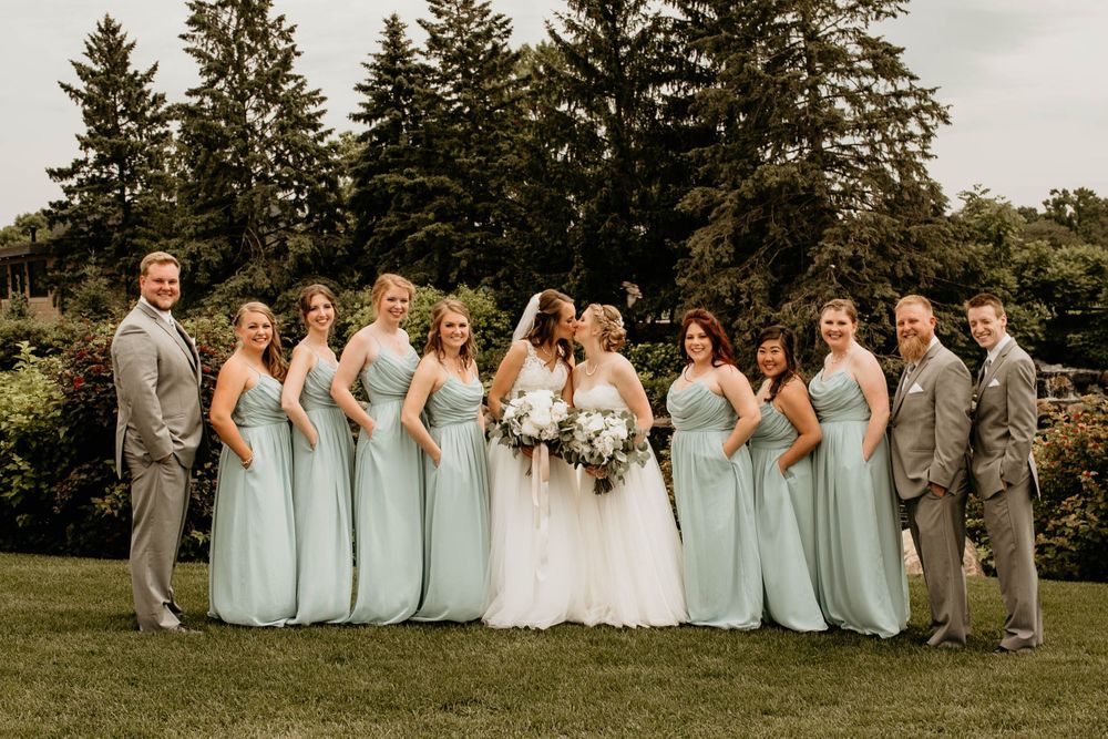 Wedding party laughs together outdoors, bridesmaids in sage green dresses flanking two brides in white gowns.