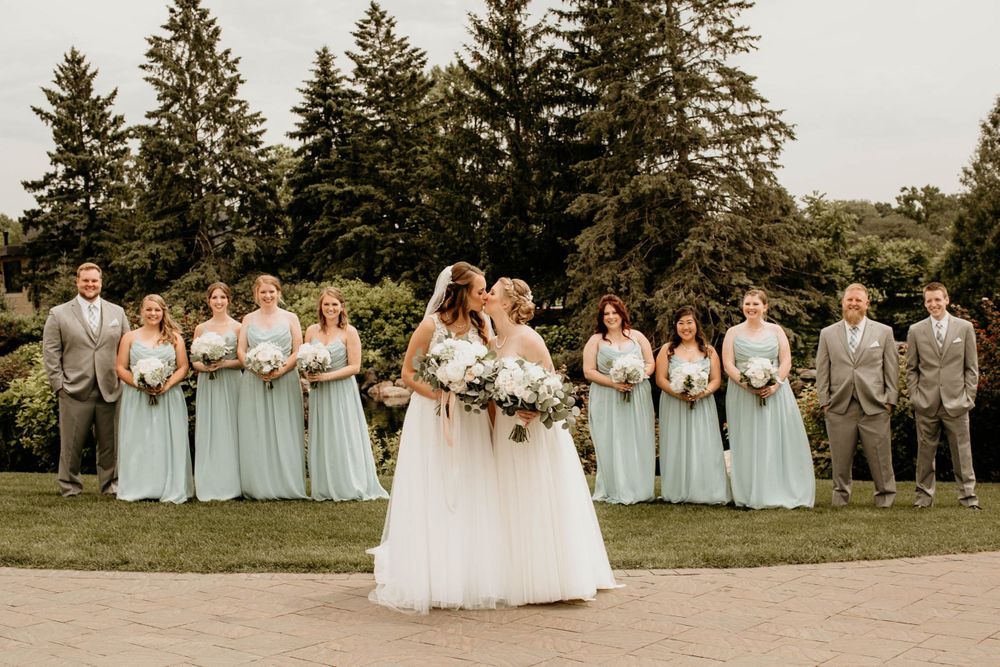 Two brides share an intimate moment as their wedding party in mint green and grey stands behind them outdoors.