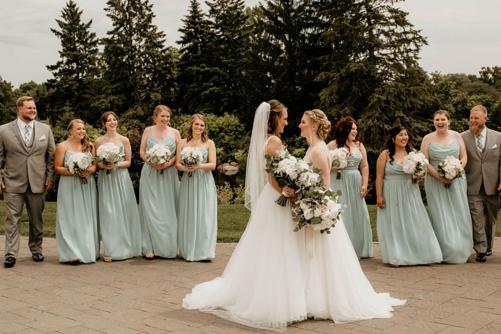 Two brides face each other holding bouquets while their wedding party in sage green poses behind them outdoors.