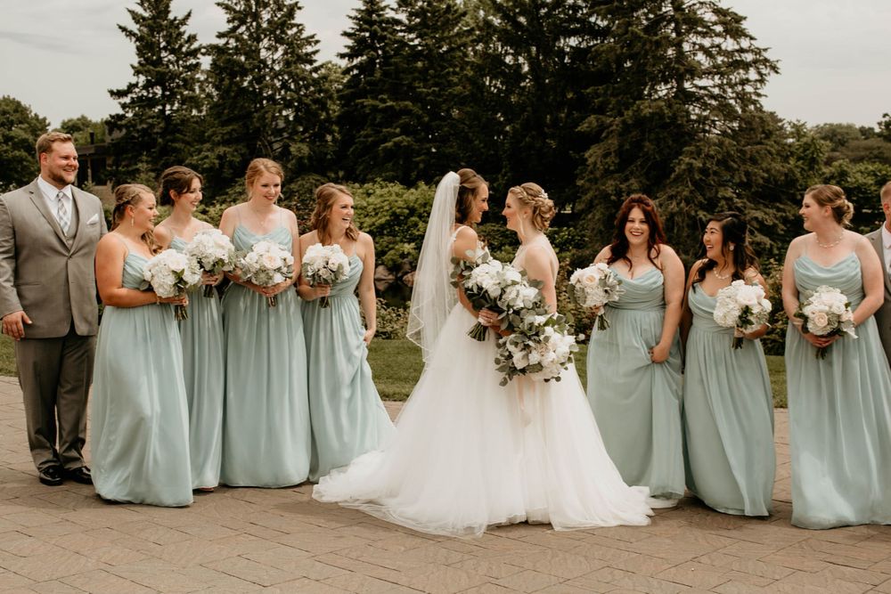 Two brides walk with bridesmaids in mint green dresses and groomsmen in grey suits through an outdoor garden setting.