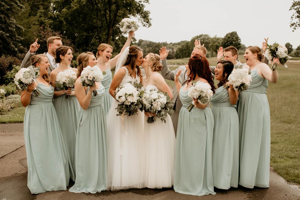 Bridesmaids in sage green cheer as two brides share a kiss, holding white floral bouquets outdoors.